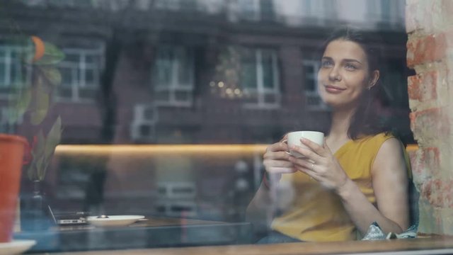 Happy young lady is drinking coffee and looking out of window sitting in cozy cafe alone smiling relaxing. Happiness, drinks and modern lifestyle concept.