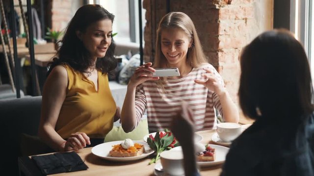 Young women happy friends are taking photo of meals in cafe shooting with smartphone camera laughing using modern gadgets. Millennials and lifestyle concept.