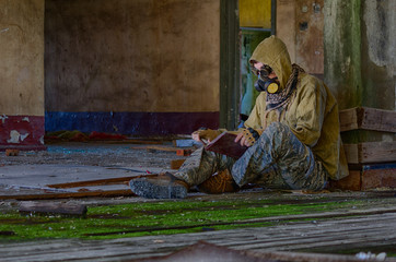 Man in respirator gas mask, cyberpunk goggles, hood and camo pants sits on the floor inside of abandoned building and reading book. Cyberpunk postapocalypse fantasy horror scene, or air pollution