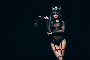 sexy young brunette woman in bdsm costume and mask with leather flogging whip isolated on black