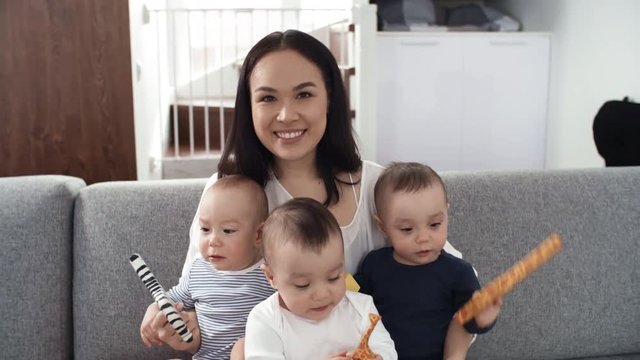 Waist-up shot of lovely middle-aged Asian mother sitting on couch at home and holding her 1-year-old identical triplet babies, who are playing and nibbling on wooden toys, looking away from camera