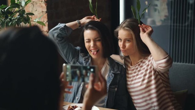 Female friends taking pictures in cafe posing with silly grimaces using smartphone touching screen enjoying leisure time together. Happiness and youth concept.