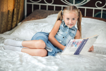 Cute little girl in denim sundress reading book looking at camera and smiling on bed at home.