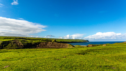 Fototapeta na wymiar Green Irish meadow landscape next to Doolin Bay with the cliff in the background, sunny day with a blue sky with white spring clouds on a calm day in County Clare in Ireland. Wild Atlantic Way