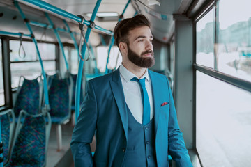 Fototapeta na wymiar Young thoughtful bearded businessman in blue suit standing in public transportation and looking trough window.