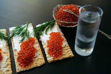 Snack with red caviar and glass of cold vodka on black wooden background. Spirits and traditional starter.
