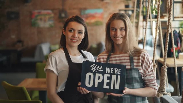 Portrait of happy girls waitresses in aprons holding we are open sign standing in cozy cafe smiling looking at camera. Modern business and people concept.