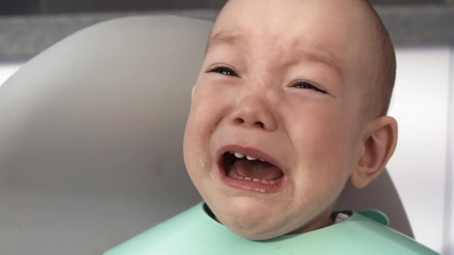 Close-up face shot of sloppy young Asian baby with large dark brown eyes, wearing bright plastic bib, getting upset and starting to cry during mealtime