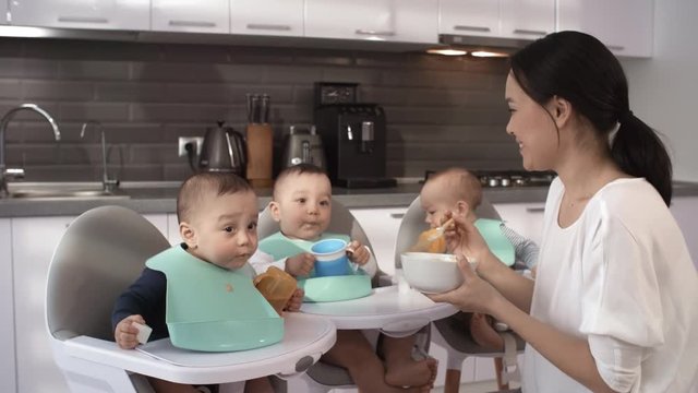 Waist-up shot of beautiful Asian mother sitting in kitchen with her 1-year-old triplet babies in high chairs, with bright bibs on necks, and feeding them puree, then turning towards camera and smiling