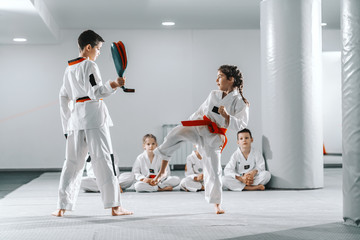Caucasain boy and girl in doboks having taekwondo training at gym. Girl kicking while boy holding kick target. In background their friend sitting with legs crossed and watching them.