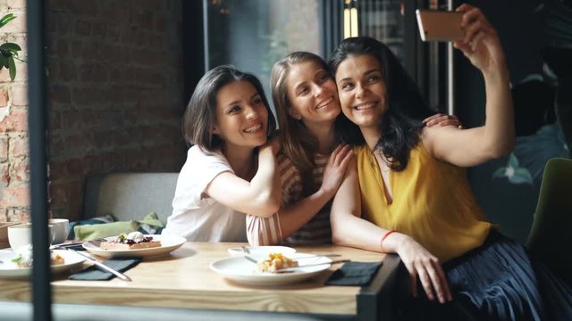 Group of friends girls taking selfie in cafe holding smartphone sitting at table having fun laughing shooting photographs. Happiness and modern technology concept.