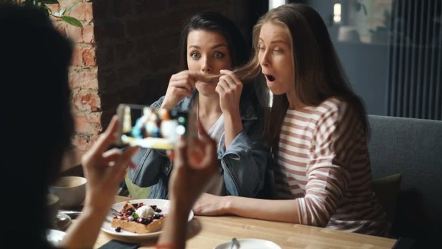 Happy young ladies playful friends are having fun in cafe taking photos with smartphone posing with hair moustache laughing. Modern lifestyle and joy concept.