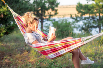 young attractive caucasian woman reading book in hammock in the forest, lake on the background