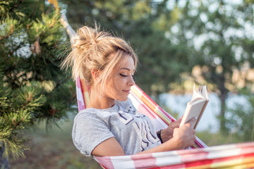 young attractive caucasian woman reading book in hammock in the forest, lake on the background