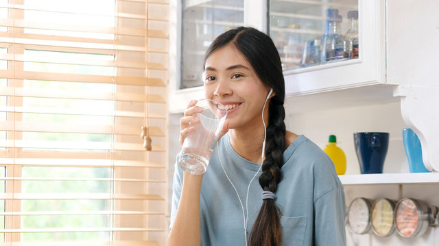 Young beautiful asian woman drinking water wearning earphones and standing by window in kitchen background, peolpe and healthy lifestyles