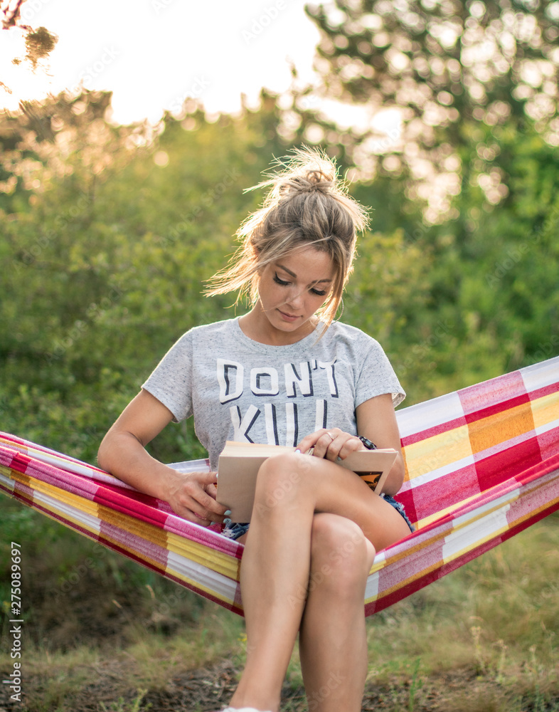 Wall mural young attractive caucasian woman reading book in hammock in the forest, sun on the background