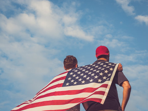 Two attractive guys holding an American Flag against a clear, sunny, blue sky. View from the back. Preparing for the holidays - Powered by Adobe