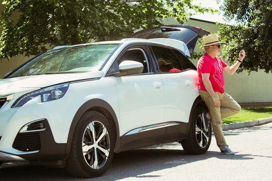 Senior man standing near by his car is working outdoors at the city's street in sunny day. Male model in sunglasses using smartphone. Concept of working, business, job, start-up, freelance.