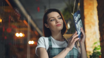 Business owner attractive woman in apron hanging we're open sign on front door smiling welcoming clients to new cafe. Business, people and youth concept.