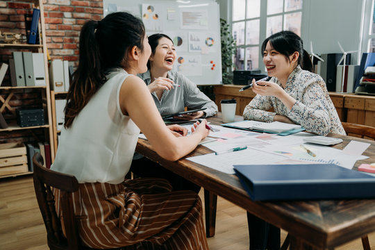 Three Asian Woman Coworkers Laughing At Funny Joke While Meeting In Office. Girls Colleagues In Friendly Work Team Enjoying Positive Emotions And Discussing Project Together. Group Of Happy Partners