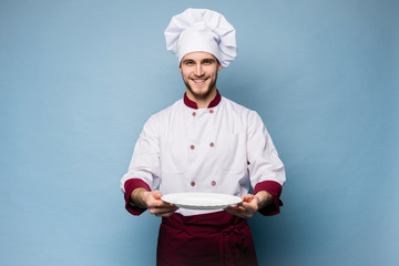 Portrait of a happy male chef cook standing with plate isolated on light blue background.