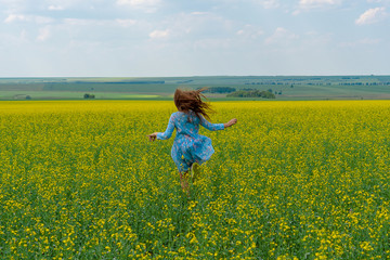 Fototapeta premium A happy girl with long hair runs across a yellow flower field. Hair develop. Yellow flowers. Concept of summer and happy childhood.