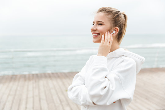 Image of happy nice woman smiling and listening to music with earpods while walking near seaside