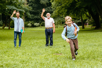 Pupil with backpack and books running at school his dad and granddad standing on background waving while seeing their little boy off to elementary school. Back to school concept.