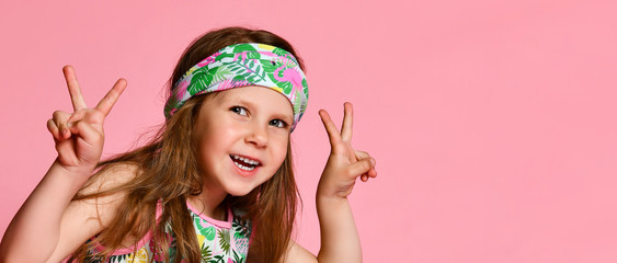 Portrait of happy cheerful girl in summer dressing for hair, showing peace gesture with two hands, isolated on pink background
