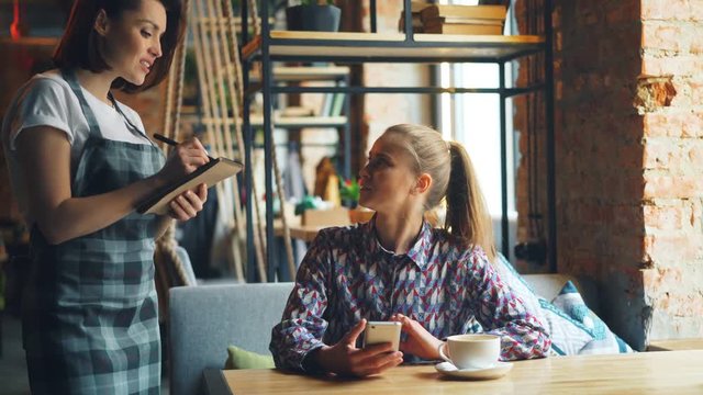 Pretty girl using smartphone in coffee house then ordering from friendly waitress talking to woman in apron smiling. Modern technology and business concept.