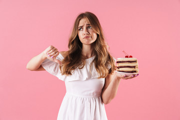 Image of disappointed sad woman wearing white dress holding piece of cake with thumb down