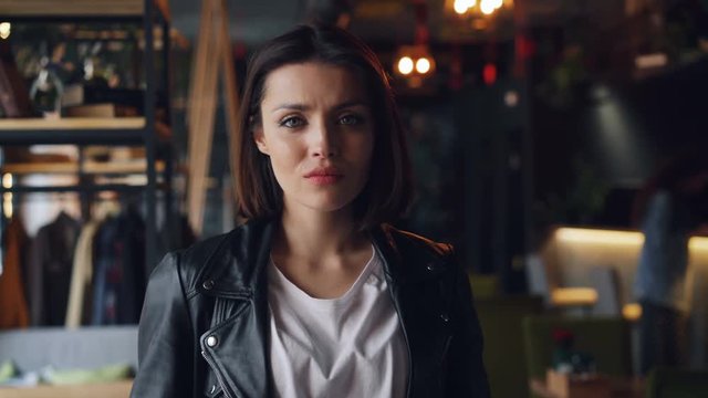 Portrait of beautiful young lady in leather jacket standing alone in cafe with serious face looking at camera. Confident people, coffee huse and youth concept.