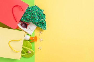 Summer concept of colourful shopping bags full of clothes. Gift bags with T-shirt, denim shorts, flip flops and bottle of sunscreen. Summer wishlist concept. Top view copy space on yellow background