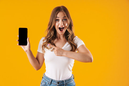 Image closeup of pretty caucasian woman wearing basic t-shirt rejoicing while pointing finger at cellphone
