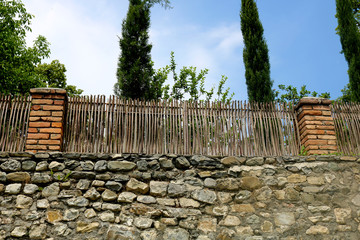 Fence of stones and thin branches. Cypresses, blue sky and fence	