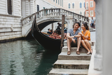 Loving couple on vacation in Venice, Italy - Millennials sitting on a canal with gondolas