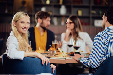 Young smiling Caucasian blonde cute woman dressed smart casual sitting at table in restaurant and looking over shoulder. In background are her friends having dinner.