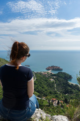 Red head girl hill enjoying Sveti Stefan island in Budva, Montenegro. Young woman looking to the adriatic sea and green cliff. Tourist traveler Relaxing watching a beach