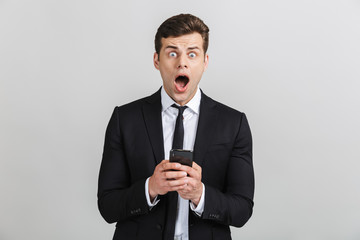 Image of shocked yong businessman in formal suit typing on cellphone and looking at camera with open mouth