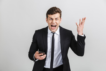 Image of angry caucasian businessman in formal suit holding cellphone and screaming at camera with hand raised