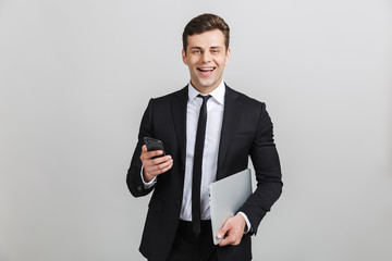 Image of young happy businessman in formal suit smiling while holding laptop and cellphone isolated