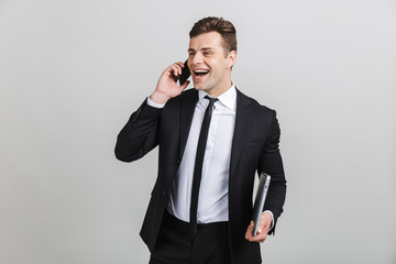 Naklejka premium Image of young cheerful businessman in formal suit talking on cellphone while holding laptop
