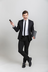 Full length image of furious businessman in formal suit screaming while holding cellphone and laptop