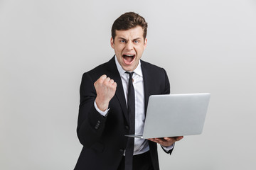 Image of successful excited businessman in formal suit celebrating triumph while holding laptop isolated over gray background