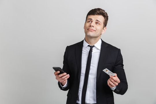 Image of thoughtful brunette businessman in formal suit dreaming while holding cellphone and credit card