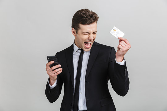 Image of delighted brunette businessman in formal suit screaming while holding cellphone and credit card