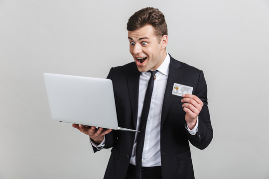 Image of amazed brunette businessman in formal suit surprising while holding laptop and credit card