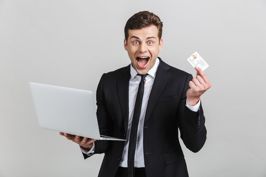 Image of ecstatic brunette businessman in formal suit surprising while holding laptop and credit card