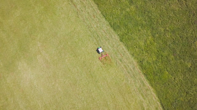 Tractor Raking Grass In An Aerial View. Tracking Shot Of Farmer Doing Field Work With Dry Hay On A Sunny Day On Meadow.