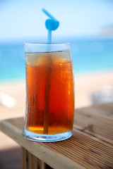 A big glass of refreshing cold ice tea is on the wooden table  with the view of unfocused beach and the blue sea 